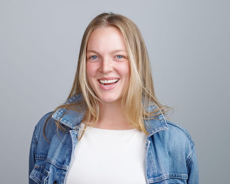 Headshot of Emma Larson laughing, in a denim jacket over a white top against a grey background