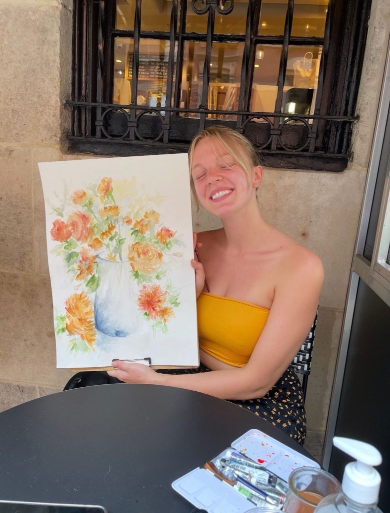 Emma Larson smiling while holding up a watercolor painting of flowers at an outdoor cafe table